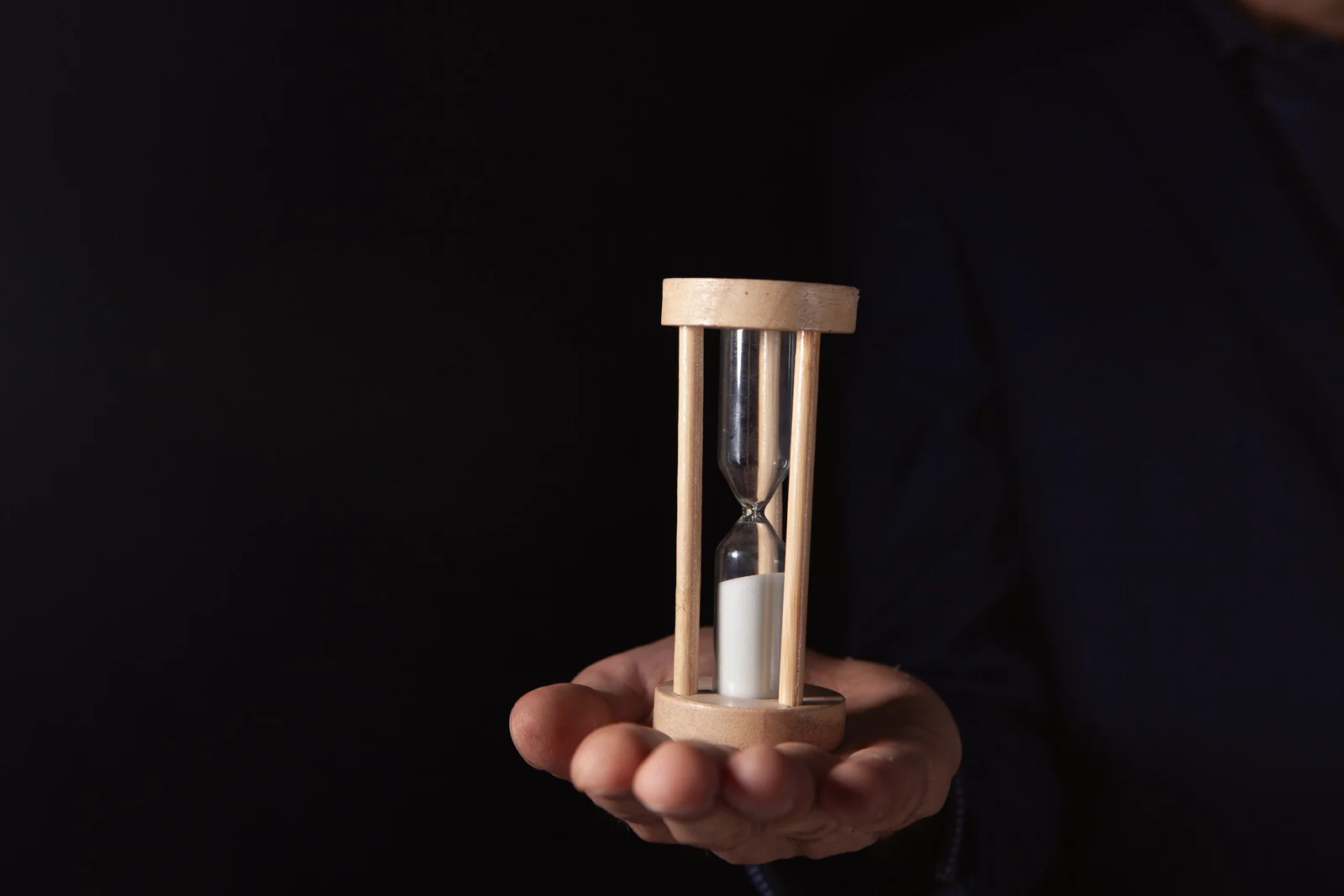 A hand holds a small wooden hourglass, sand running into the lower glass, against a black background.