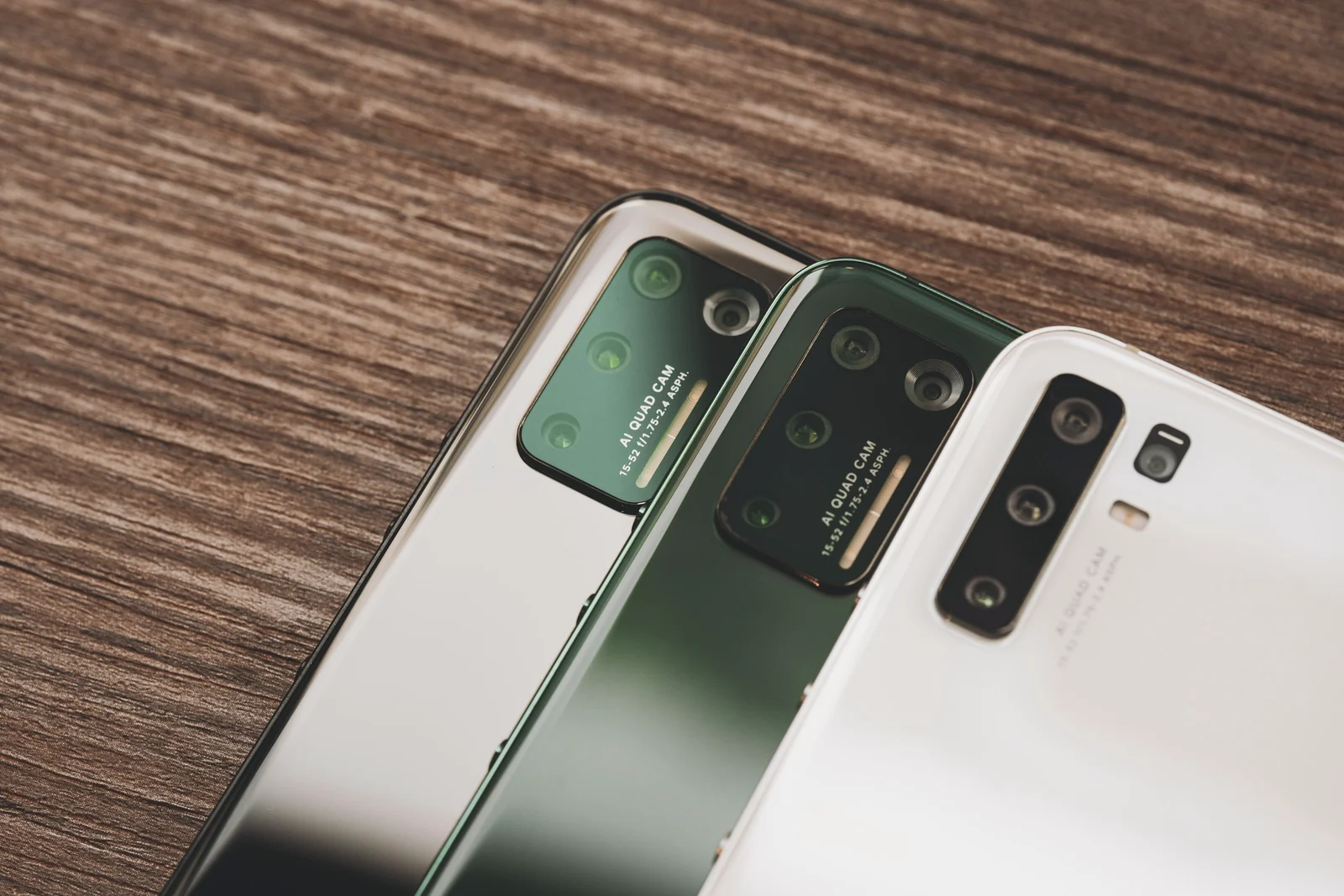 Three phones lie face down side by side on a warm wooden table, their camera bumps catching the light.